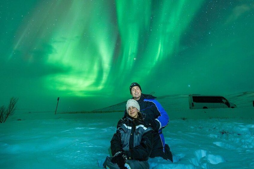 Two people posing under the Northern Lights with a snow-covered landscape and a van in the background. 1
