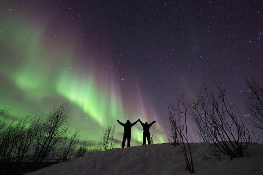 Two people standing on a snowy hill with arms raised, under a night sky with the aurora borealis. 2
