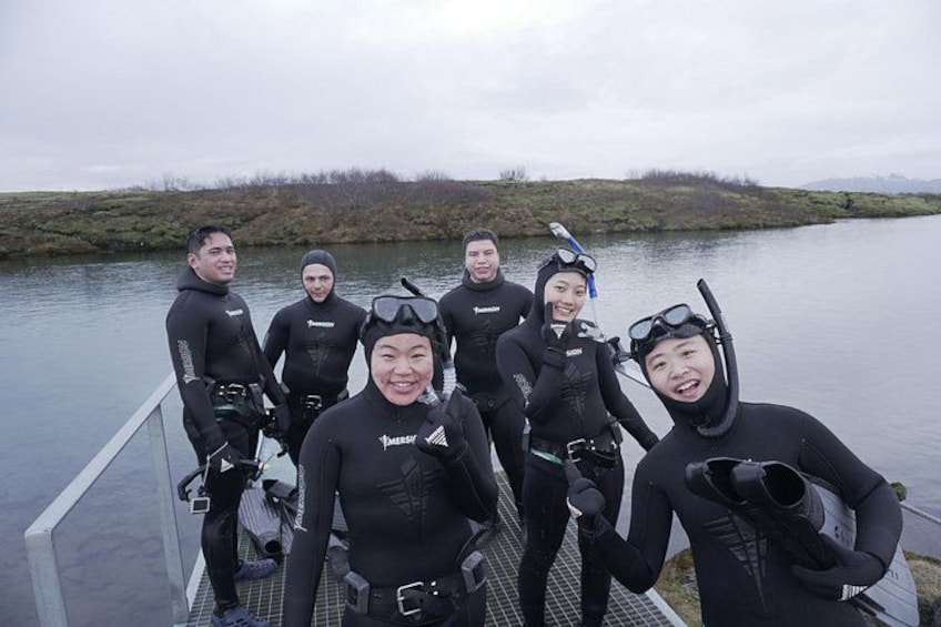 A group of people in black wetsuits standing on a dock by a body of water. 1