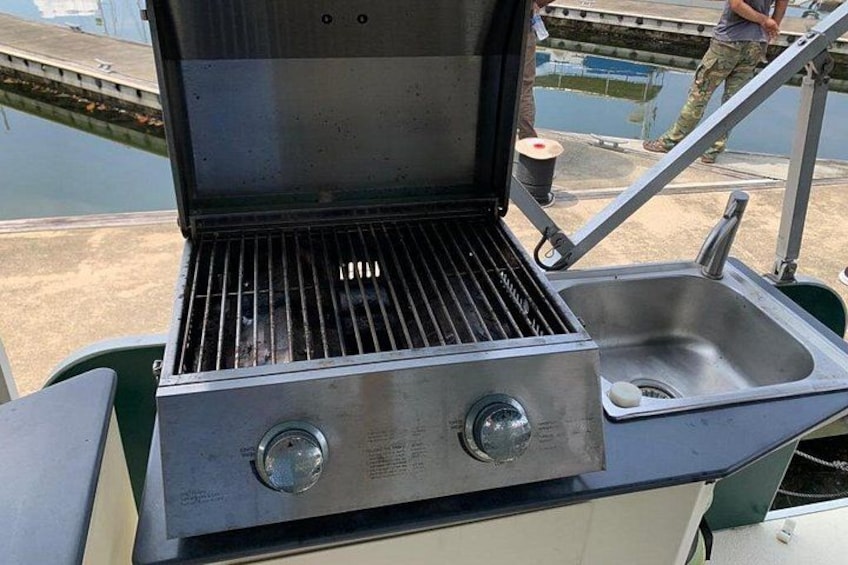 Ein tragbarer Grill mit Spüle und zwei Drehknöpfen, draußen in der Nähe eines Stegs platziert. 3