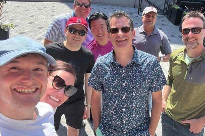 A group of people standing outdoors, some wearing hats and sunglasses, with a paved surface and potted plants in the background. 4