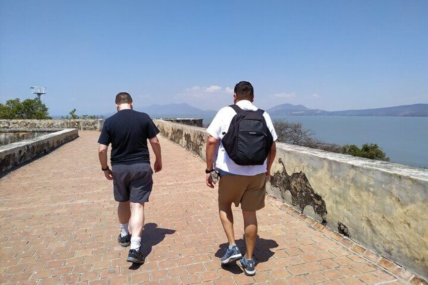 Two men walking on a brick pathway by a body of water with mountains in the background. 3