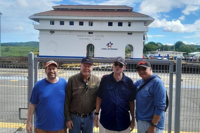 Four men standing in front of a building with the text 'MIRAFLORES LOCKS CANAL DE PANAMA' and a lock symbol. 4