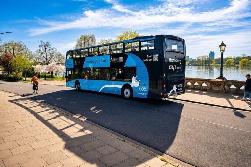 A double-decker bus with 'Hamburg City Tours' branding, parked on a paved area near a body of water with trees and a lamp post in the background. 3