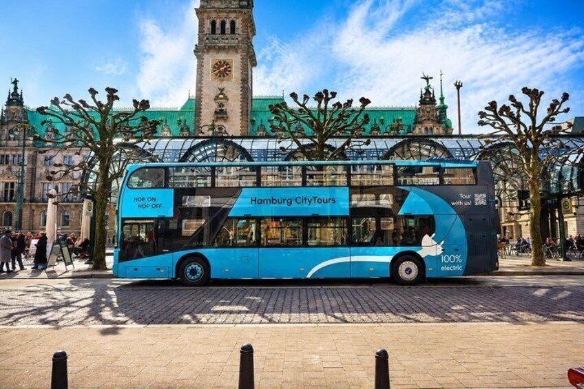 A blue double-decker bus with 'Hamburg CityTours' and '100% electric' on the side, parked in front of a historic building with a clock tower. 1