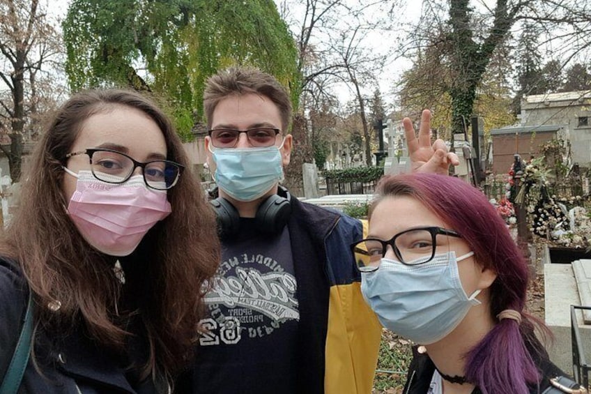Trois personnes prennent un selfie dans un cimetière, avec des pierres tombales et des arbres en arrière-plan. 2