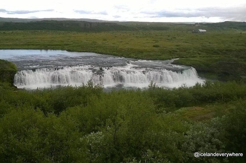 Ein Wasserfall in einer grünen Landschaft, im Hintergrund ein Haus. 2