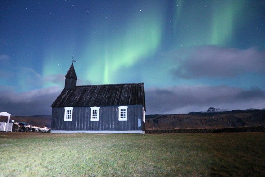 Een houten huis met een torenspits, voor een achtergrond van een nachthemel met het Noorderlicht. 5