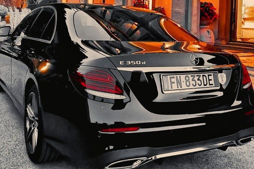 En sort Mercedes-Benz E-klasse sedan med synlig nummerplade FN 833DE, parkeret i et bymiljø med bygninger og en mur i baggrunden. 3