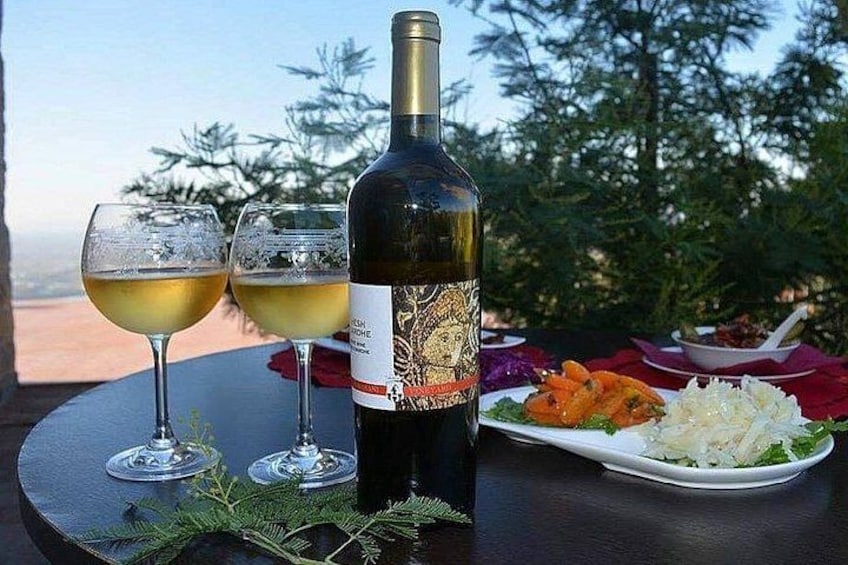 Una botella de vino, dos copas de vino y un plato de comida sobre una mesa al aire libre, con árboles al fondo. 2