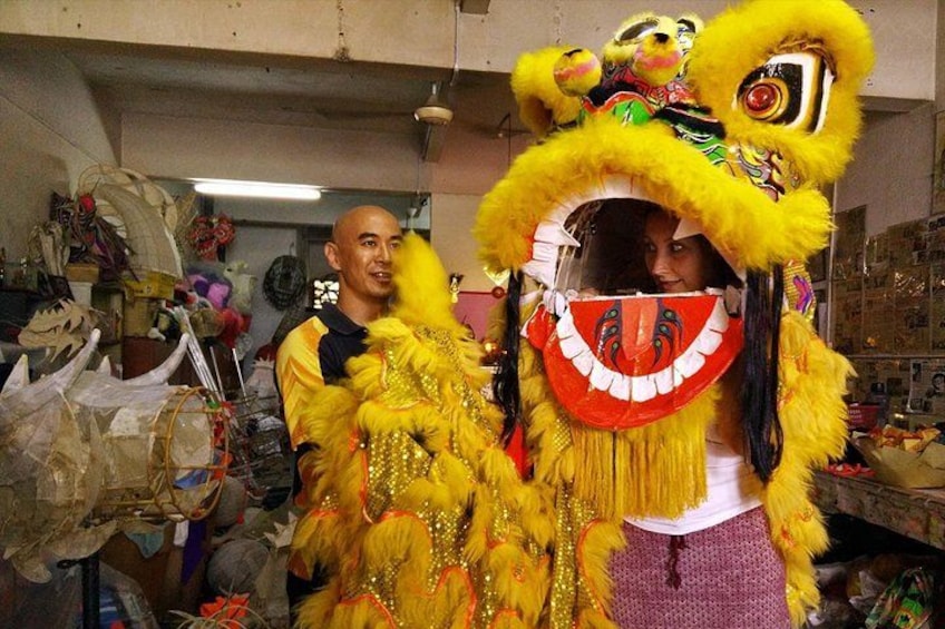 A person in a yellow lion dance costume with a red mouthpiece, standing in a cluttered room with various items and decorations. 1