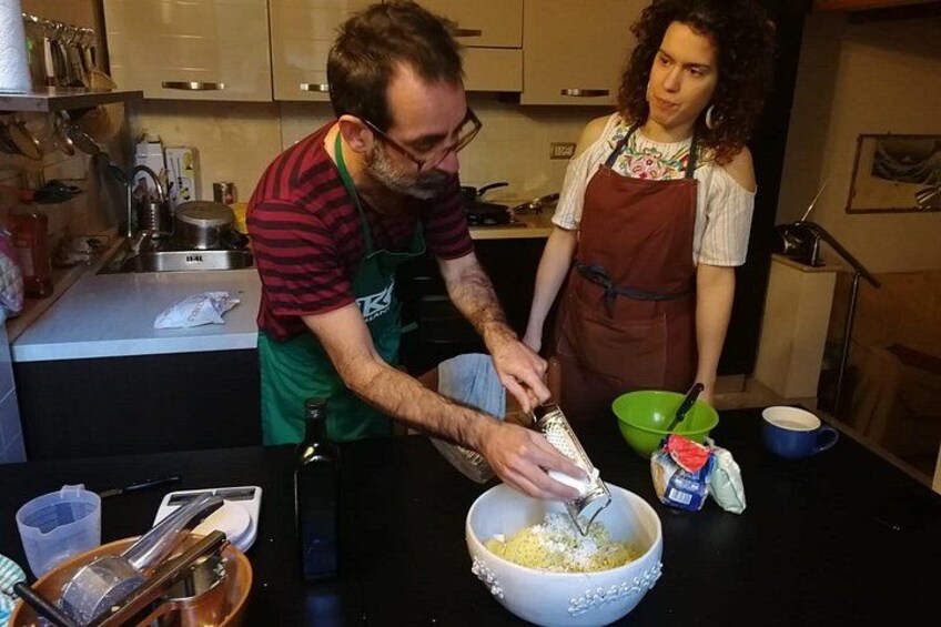 En person river ost i en skål, en annan person står bredvid, och köksredskap och ingredienser ligger på bänkskivan. 5