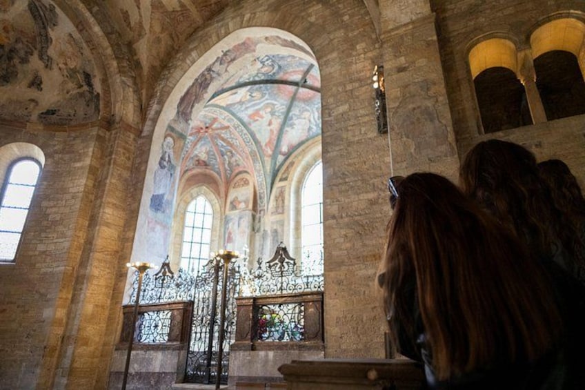 An interior of a church with arched windows, a stained glass window, and a mural on the ceiling. 5