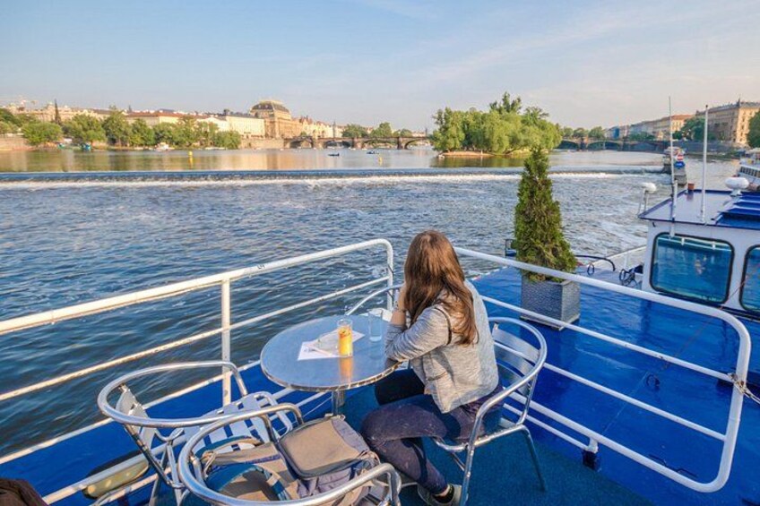 Una persona seduta a un tavolo su una barca, con un fiume e uno skyline urbano sullo sfondo. 2