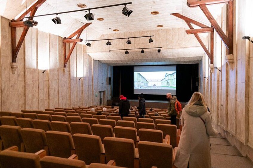 An indoor auditorium with rows of seats, a stage with a projection screen, and wooden beams on the ceiling. 5