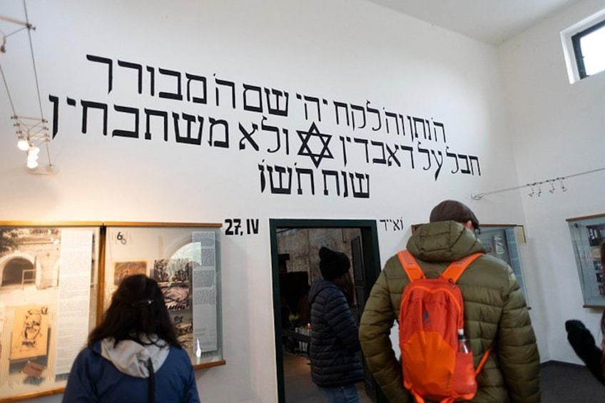 An indoor exhibition space with a wall displaying Hebrew text and a door labeled '27.IV'. 4