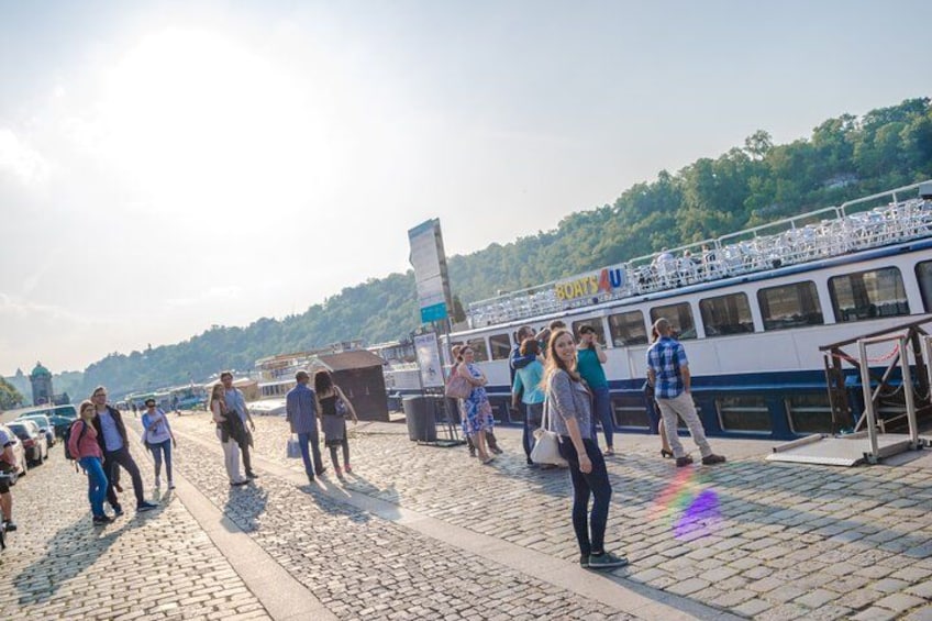 A group of people walking on a cobblestone path near a river, with a boat docked on the riverbank. 2