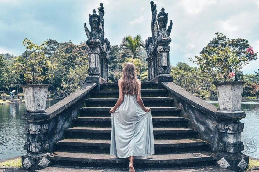 A woman in a white dress standing on stone steps with statues on either side, overlooking a body of water. 4