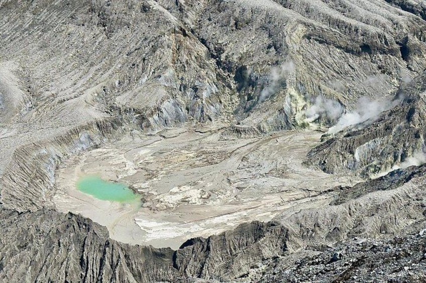 A large crater with a greenish pool, steep rocky slopes, and steam rising from the ground. 3