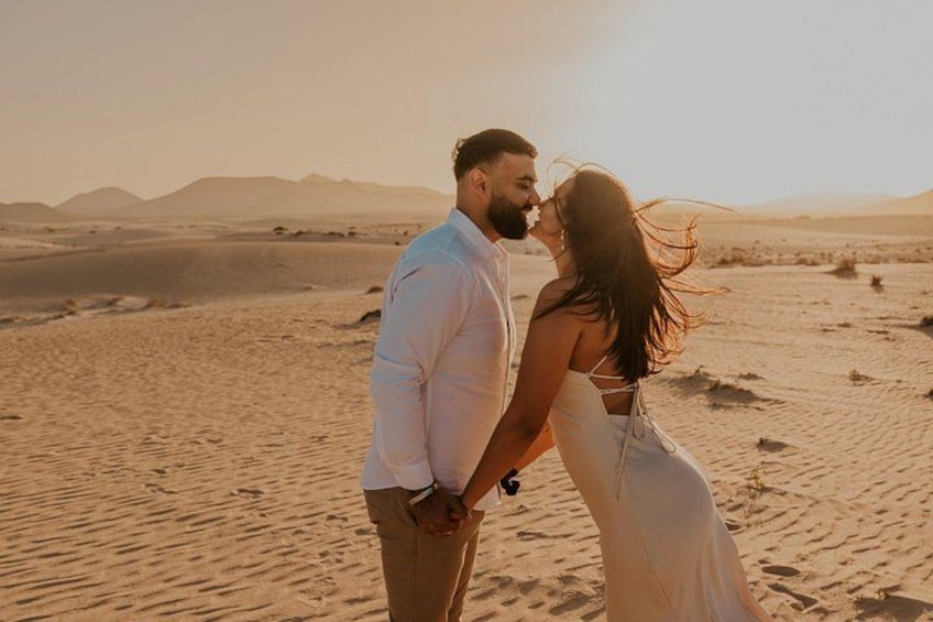 A couple embracing in a desert landscape with sand dunes and mountains in the background. 1