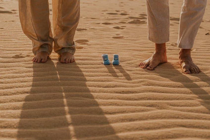 Two people standing on a sandy beach with footprints and a pair of blue baby shoes in the sand. 3