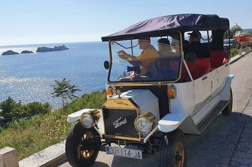 A vintage Ford car parked on a coastal road with a clear view of the sea and islands in the background. 3
