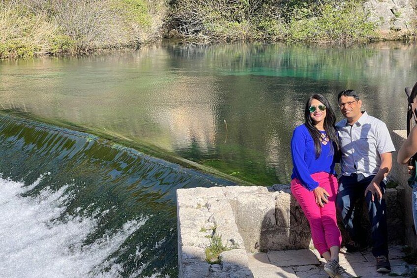 A calm river with a waterfall, people standing by the edge, and a stone wall. 2