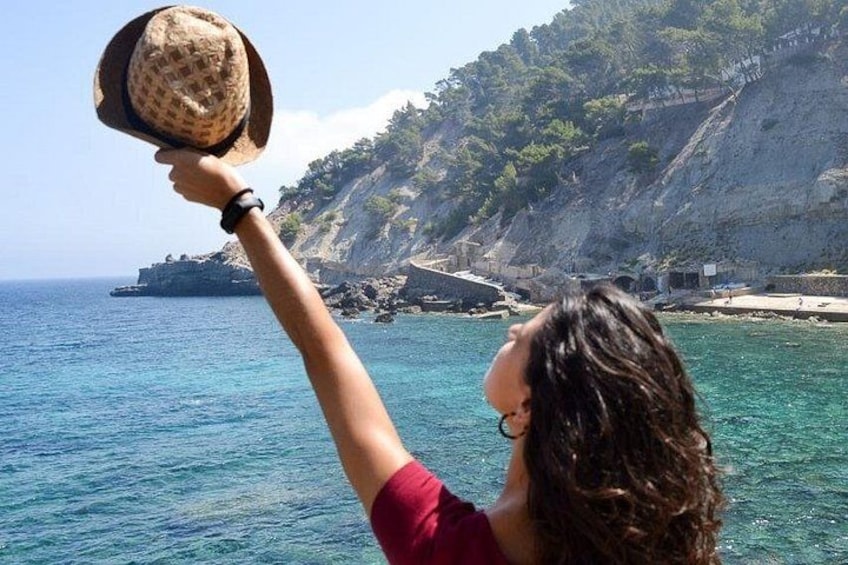A person holding a hat by the sea with a rocky coastline in the background. 1