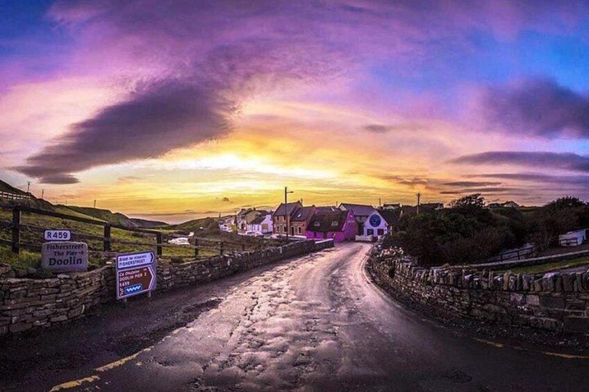 A rural road leading to a village with houses, a stone wall, and a sunset sky. 1