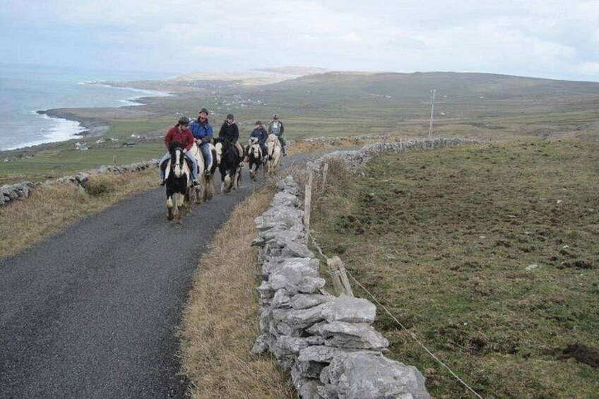 A group of people riding horses on a narrow road with a stone wall on one side and a grassy landscape with a body of water in the distance. 3