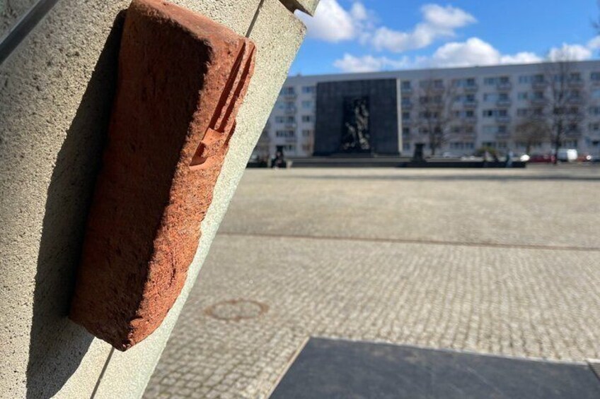 A brick leaning against a concrete wall, a paved plaza, and a building with a statue in the background. 3