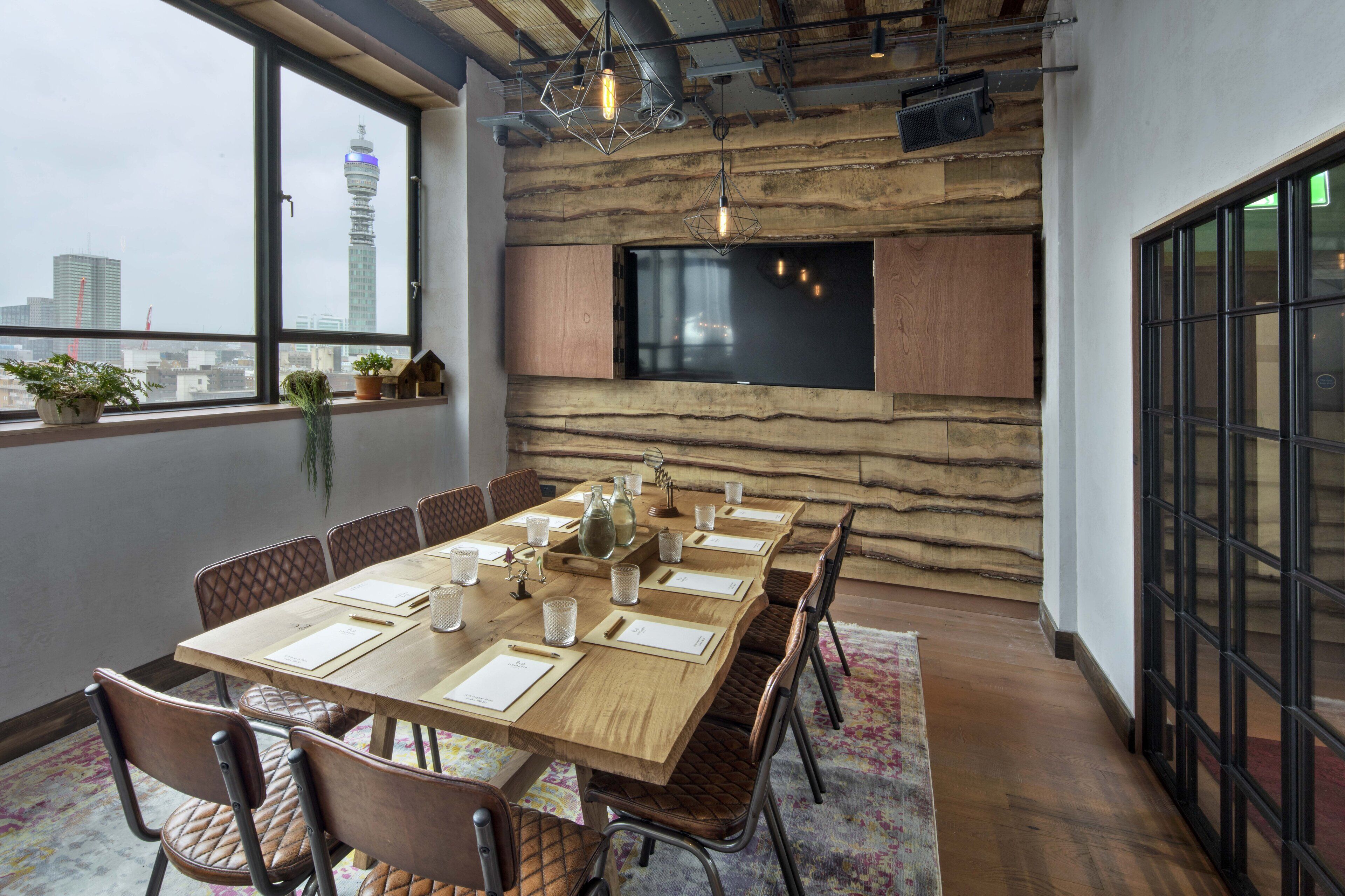 A modern dining area with a long wooden table, cushioned chairs, and a city view through large windows.