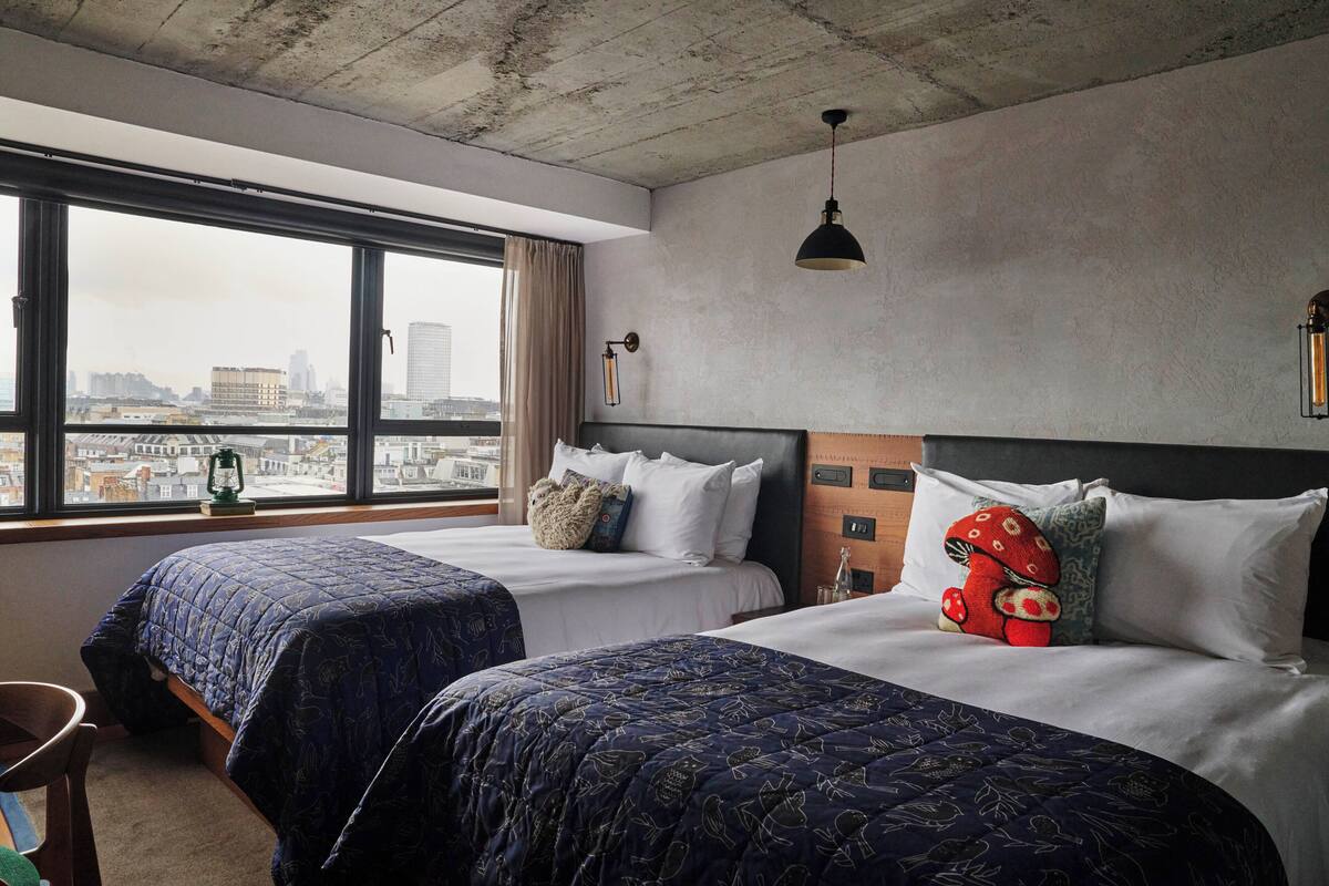 A hotel room with two beds, a large window with a city view, a hanging lamp, and a small table.