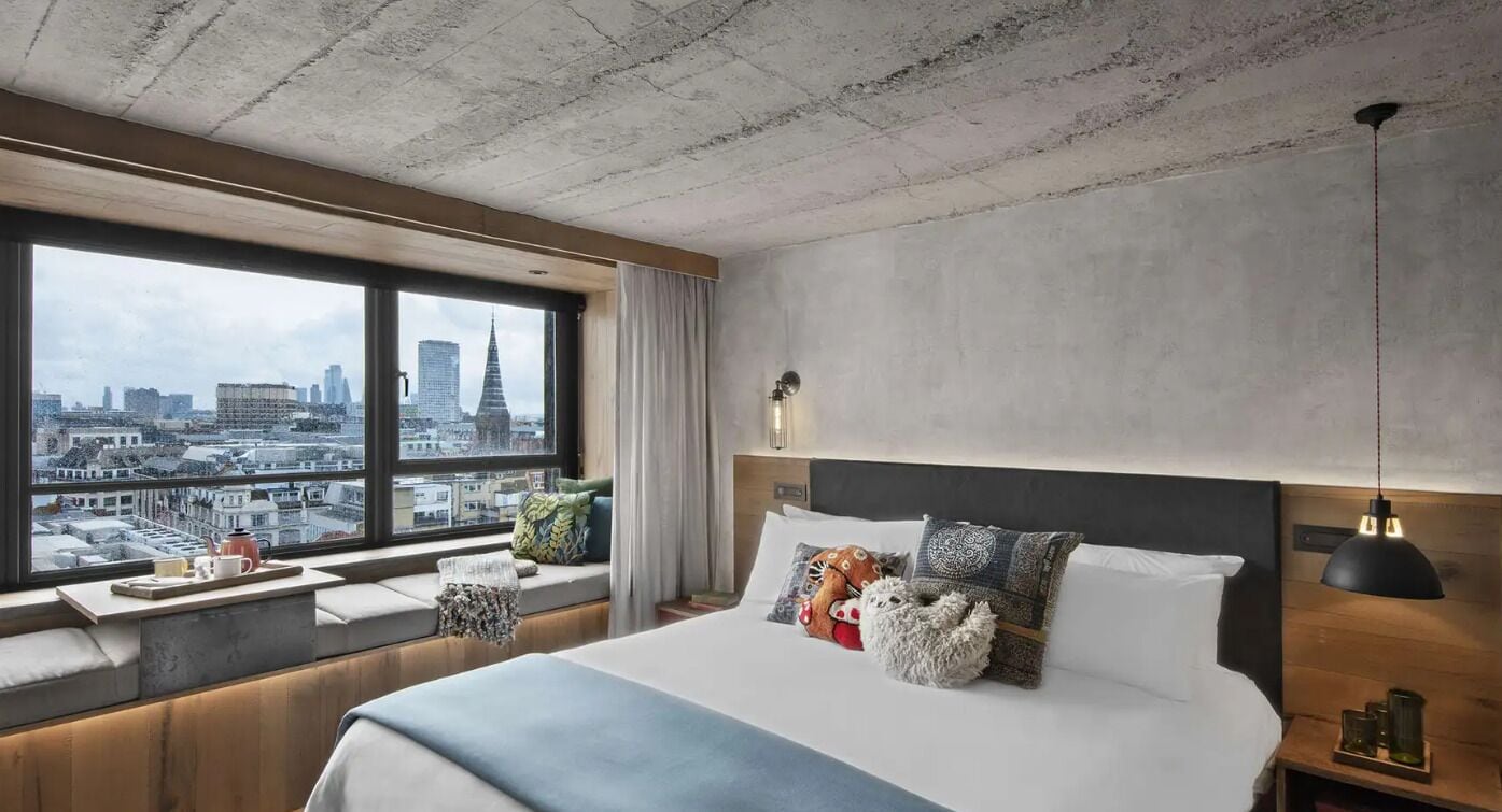 Habitación de hotel con un ventanal grande, una cama con varias almohadas, una mesita de noche con lámpara y vistas a la ciudad.