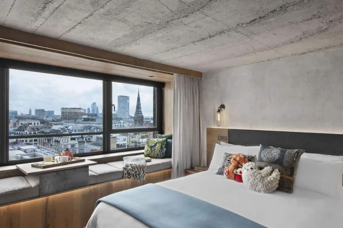 Habitación de hotel con un ventanal grande, una cama con varias almohadas, una mesita de noche con lámpara y vistas a la ciudad.