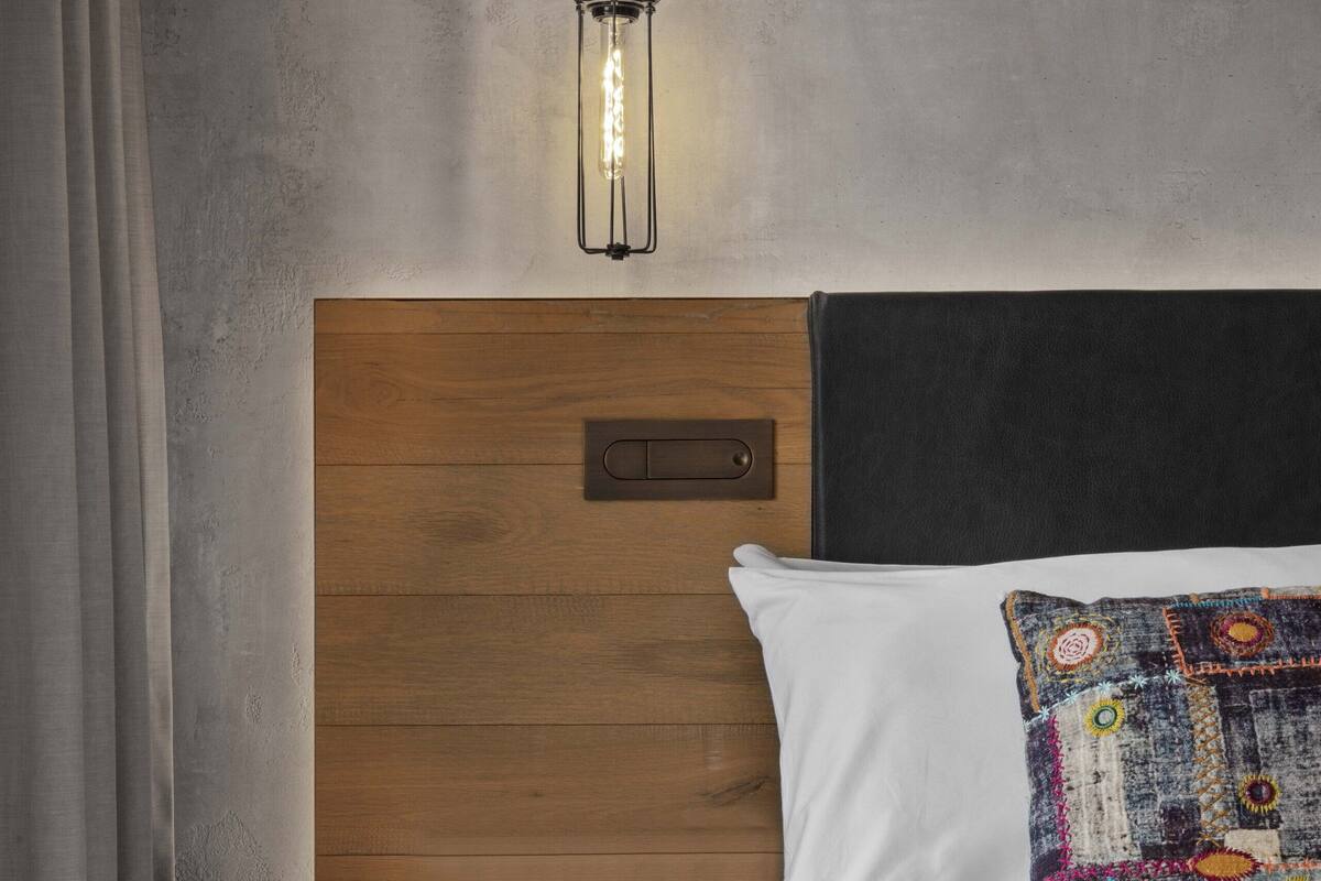 A hotel room with a wooden headboard, a bedside table with a red drawer, a lamp, and a colorful pillow.