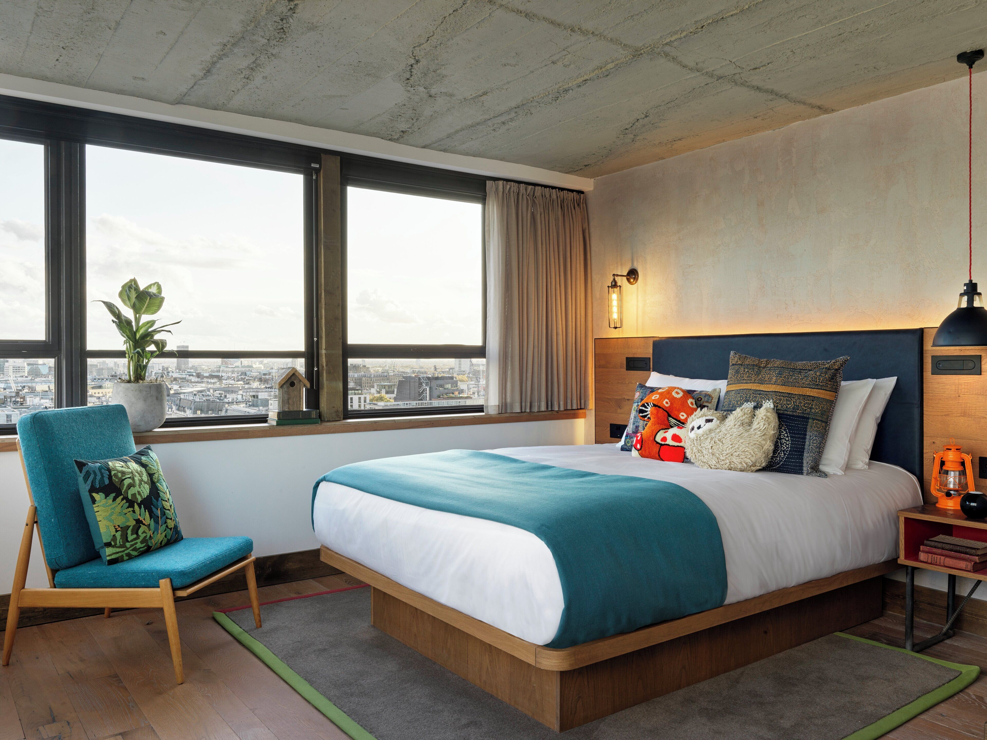 A modern hotel room with a large bed, a view of the city, a chair, and a nightstand with a lantern.