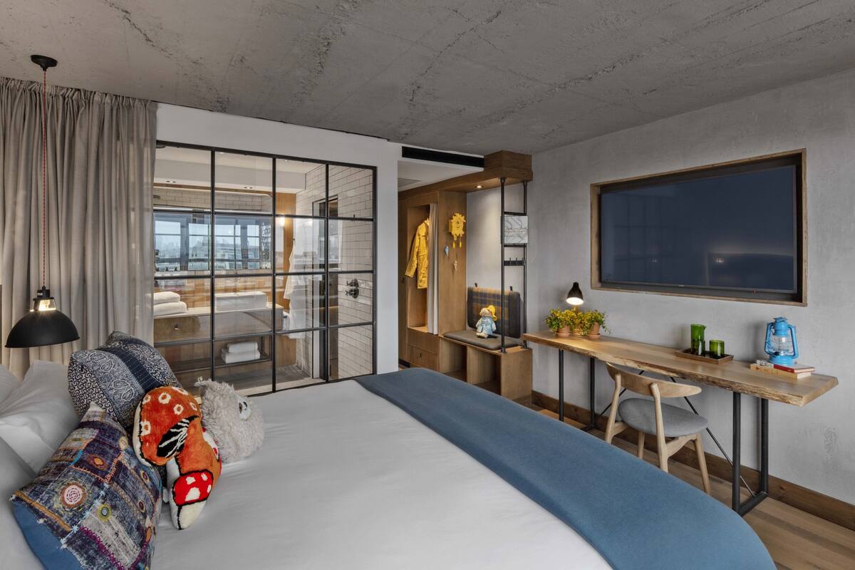 A modern hotel room with a large bed, a desk, and a view through a glass wall.