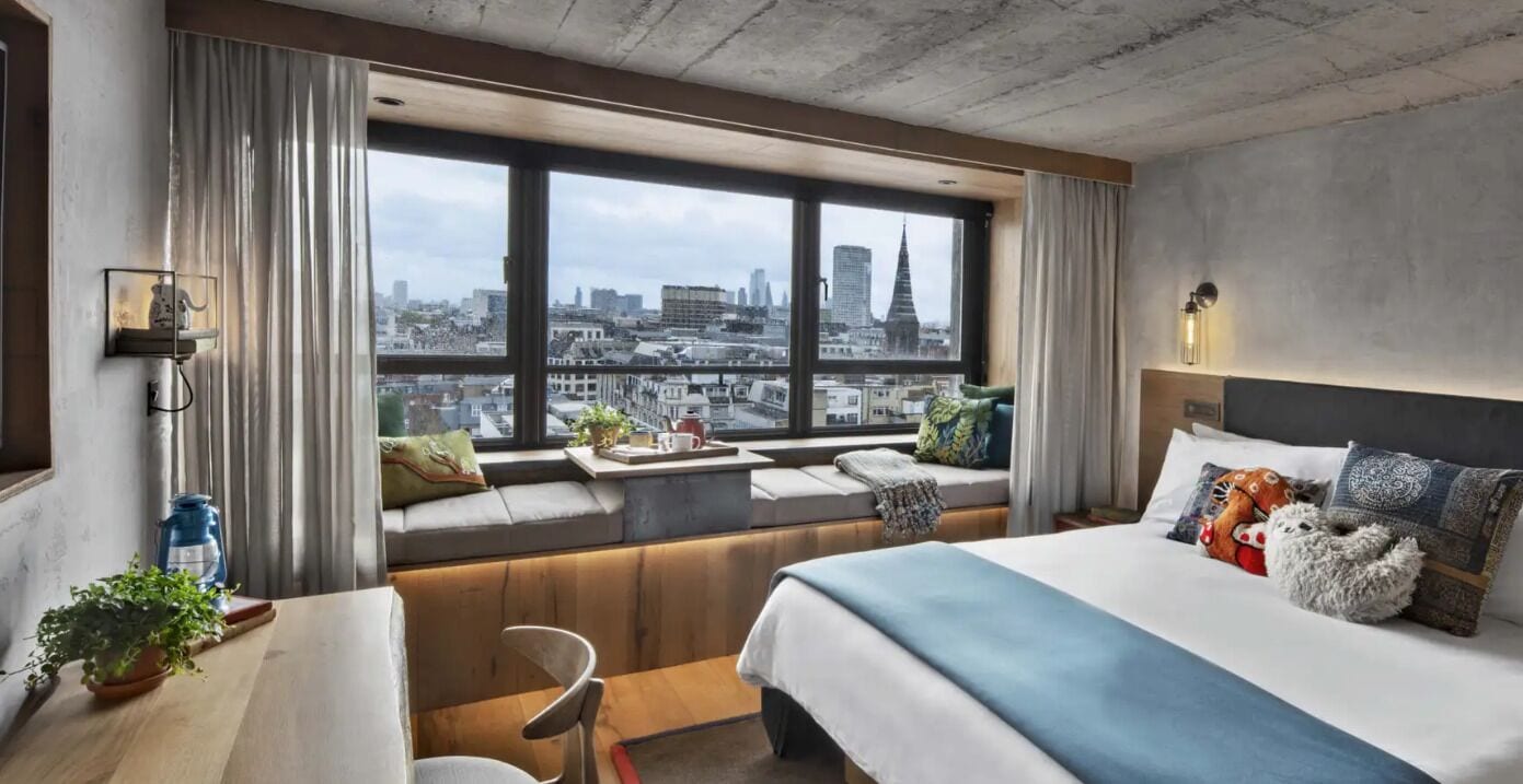Habitación de hotel con un ventanal grande, una cama, zona de estar y una mesa con una planta.