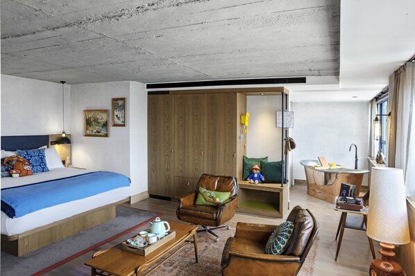A modern hotel room with a bed, a leather armchair, a sofa, a wooden cabinet, and a small bathroom.