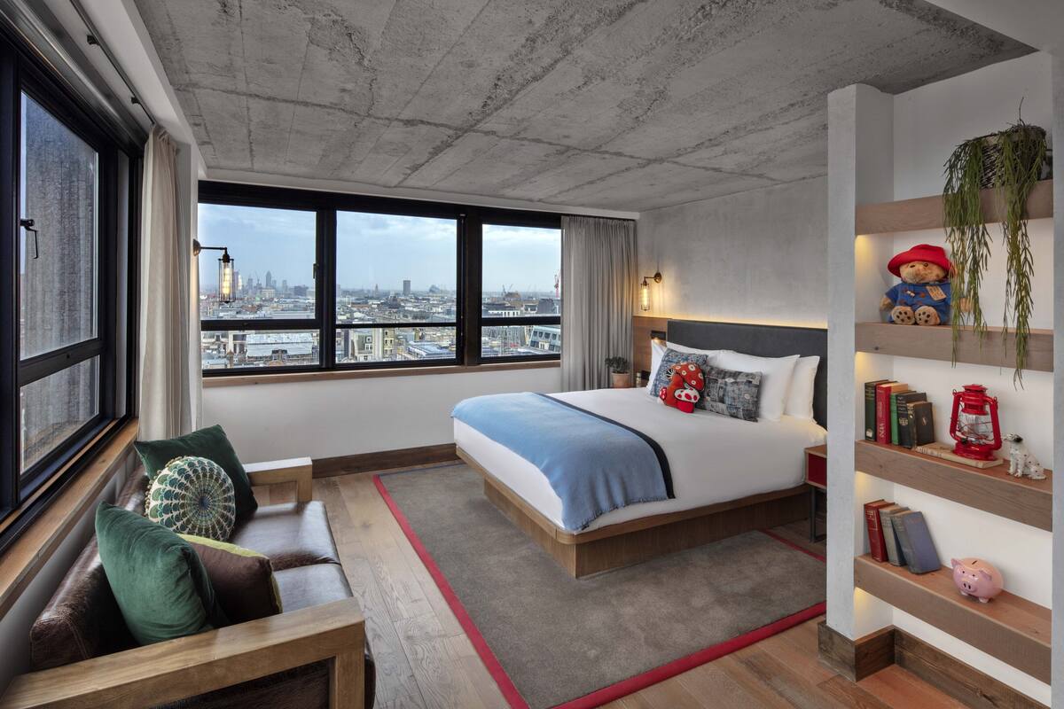 A modern bedroom with a large bed, a view of the city, a seating area, and a shelf with decorative items.