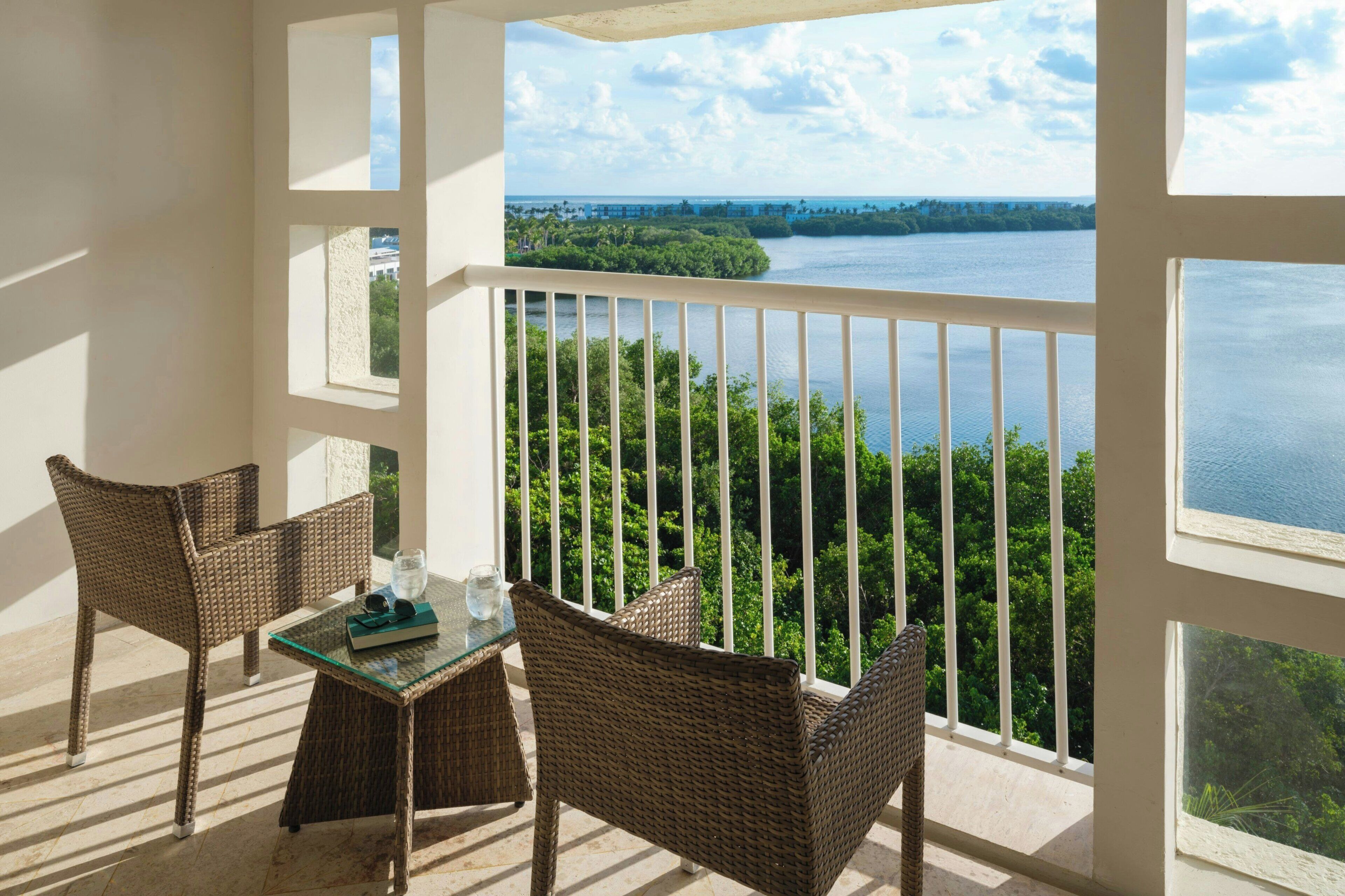 A balcony with wicker furniture, a glass table, and a view of a body of water and greenery.