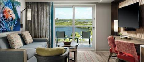 A modern living room with a sofa, chairs, a dining table, and a TV. A large window offers a view of a body of water and greenery.