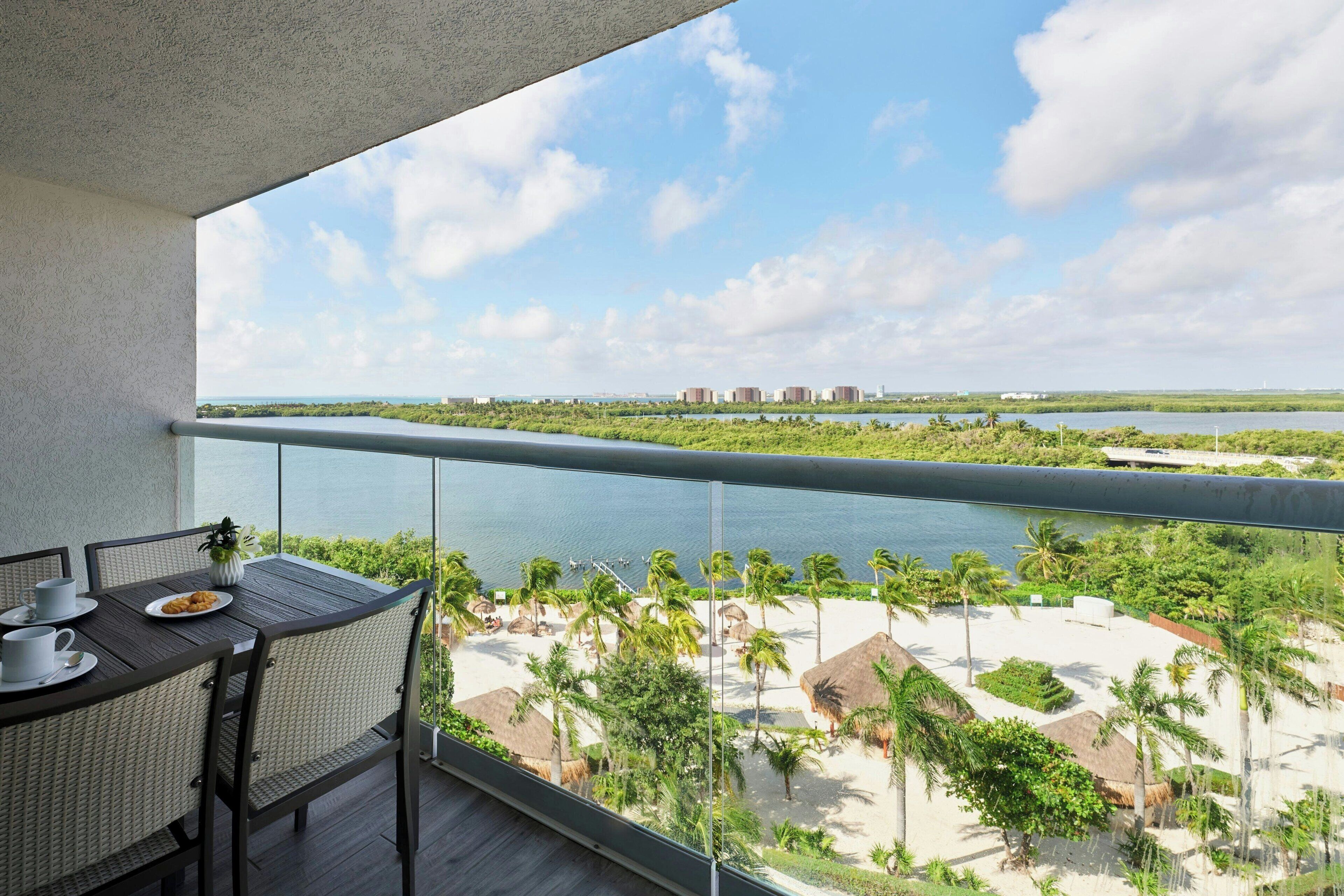 Een balkon met uitzicht op een strand en water, een eettafel met stoelen, en een kleine plant.