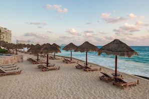 A beach with rows of sun loungers and thatched umbrellas, a pool, and a view of the ocean and distant buildings. - The Westin Cancun Resort & Spa (Cancun)