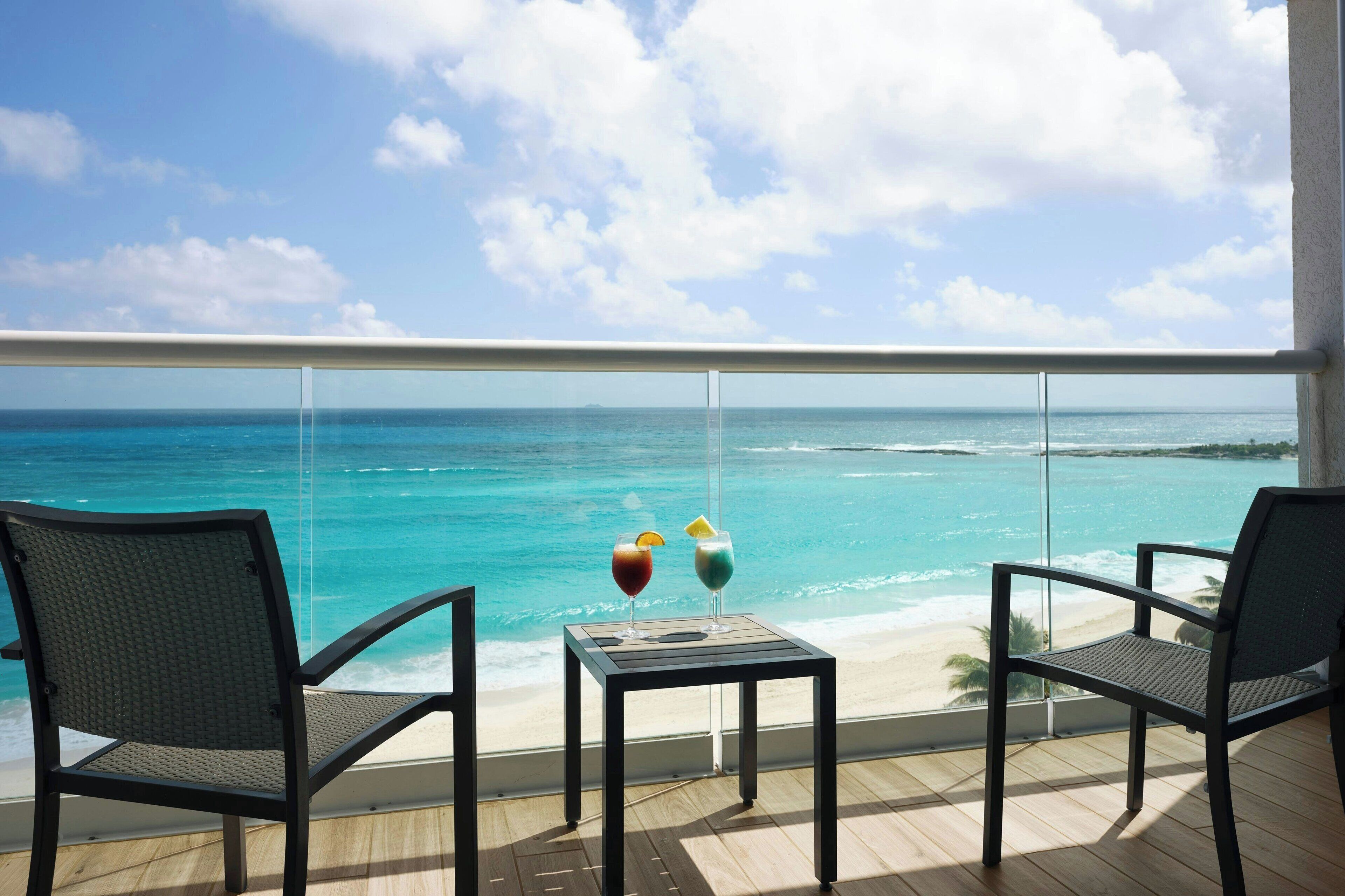 Ein Balkon mit zwei Stühlen und einem kleinen Tisch, der Blick auf einen Strand mit türkisblauem Wasser und einem klaren Himmel bietet.