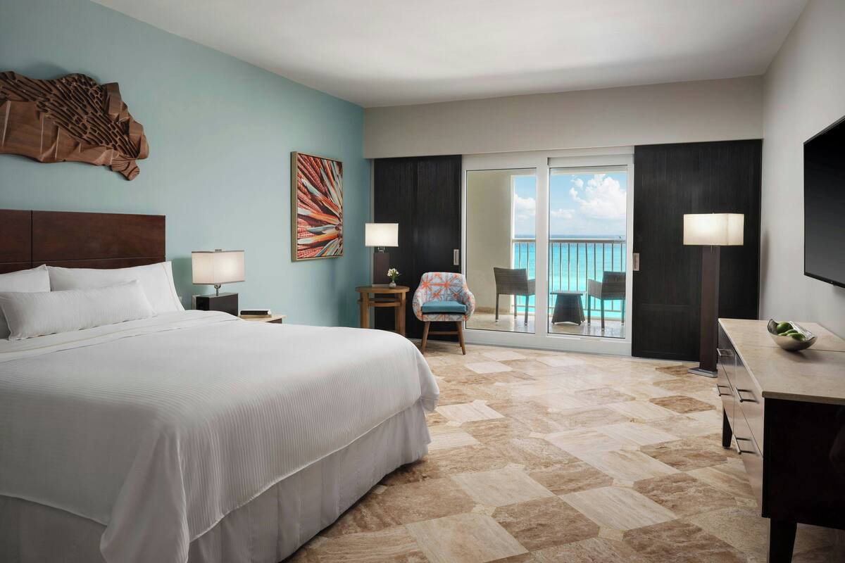 A hotel room with a large bed, a desk, a chair, a balcony with a view, and a TV.