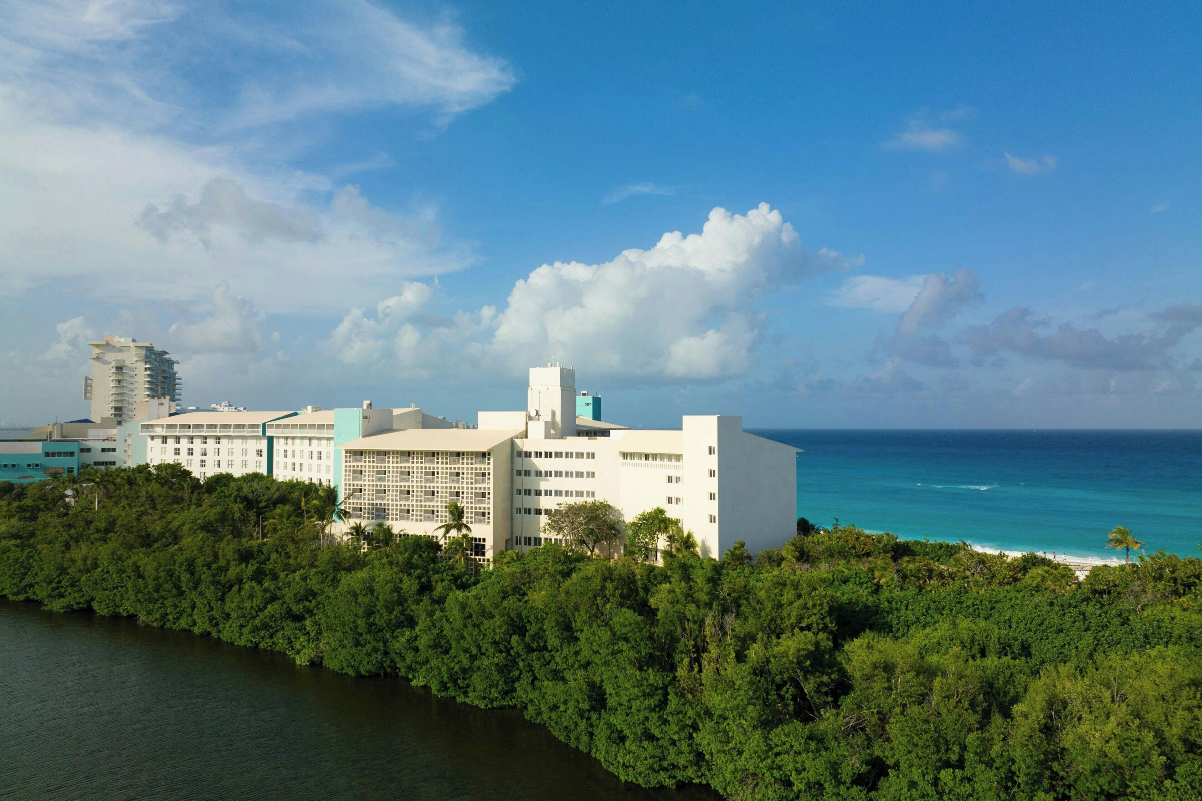 A coastal hotel with multiple buildings, a beach, and a dense forest in the foreground.