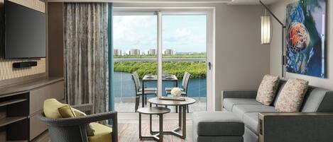 A modern hotel room with a flat-screen TV, a grey sofa, a wicker chair, a dining table, and a view of a cityscape through large windows.