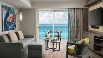 A modern living room with a sofa, armchair, dining table, and chairs, a flat-screen TV, and a large window with a view of the ocean.